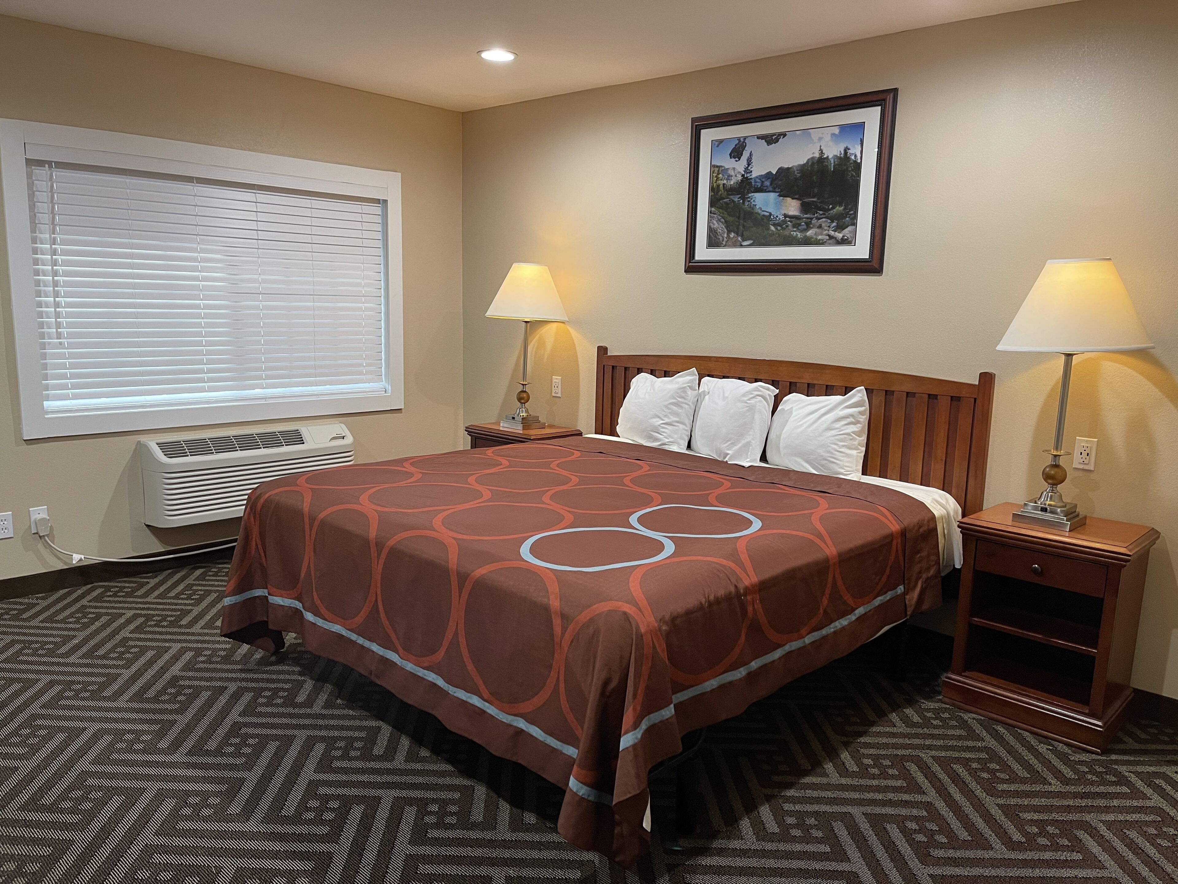 A hotel room with a large bed, two bedside lamps, a window with blinds, a wall-mounted air conditioner, and a framed picture on the wall.