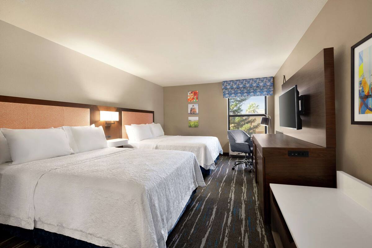 A hotel room with two beds, a desk, a chair, a TV, and a window with a view.