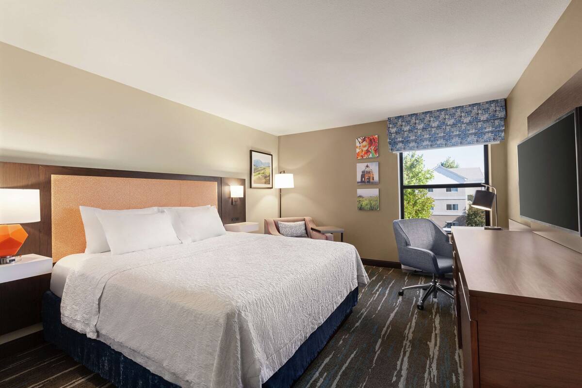 A hotel room with a large bed, a desk, a chair, and a window with a view of buildings.
