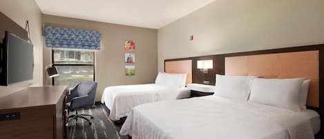 A hotel room with two beds, a desk, a chair, a television, and a window with a view of greenery.