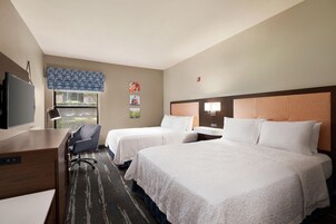 Quarto de hotel com duas camas, uma escrivaninha, uma cadeira, uma televisão e uma janela com vista para o verde.