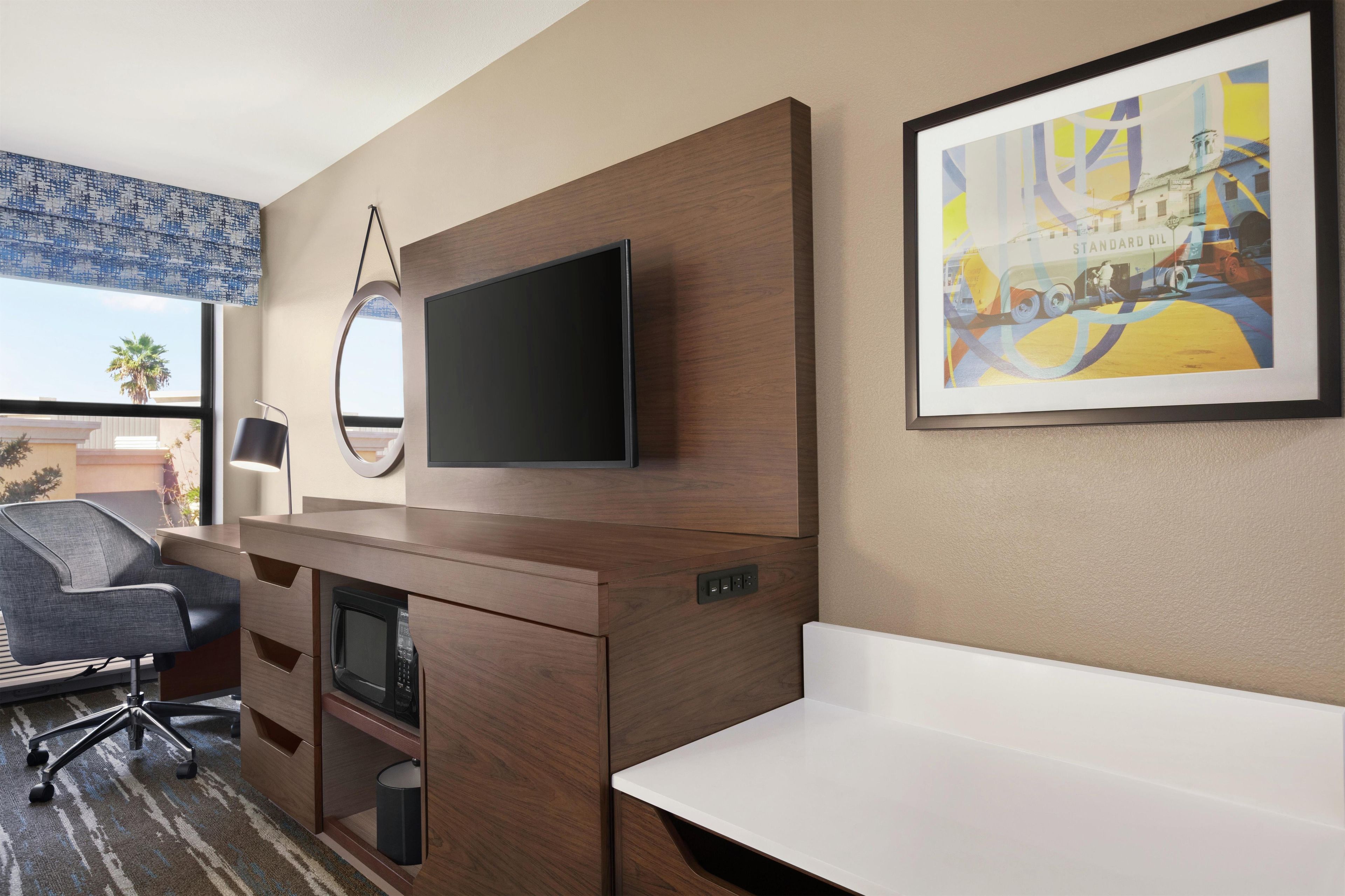 Habitación de hotel con televisión de pantalla plana montada en la pared, un escritorio con una silla, un microondas y un pequeño bote de basura.