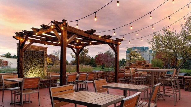 An outdoor patio area with wooden tables and chairs, a stone wall, and string lights.