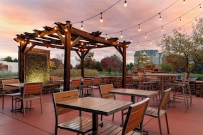 An outdoor patio area with wooden tables and chairs, a stone wall, and string lights.