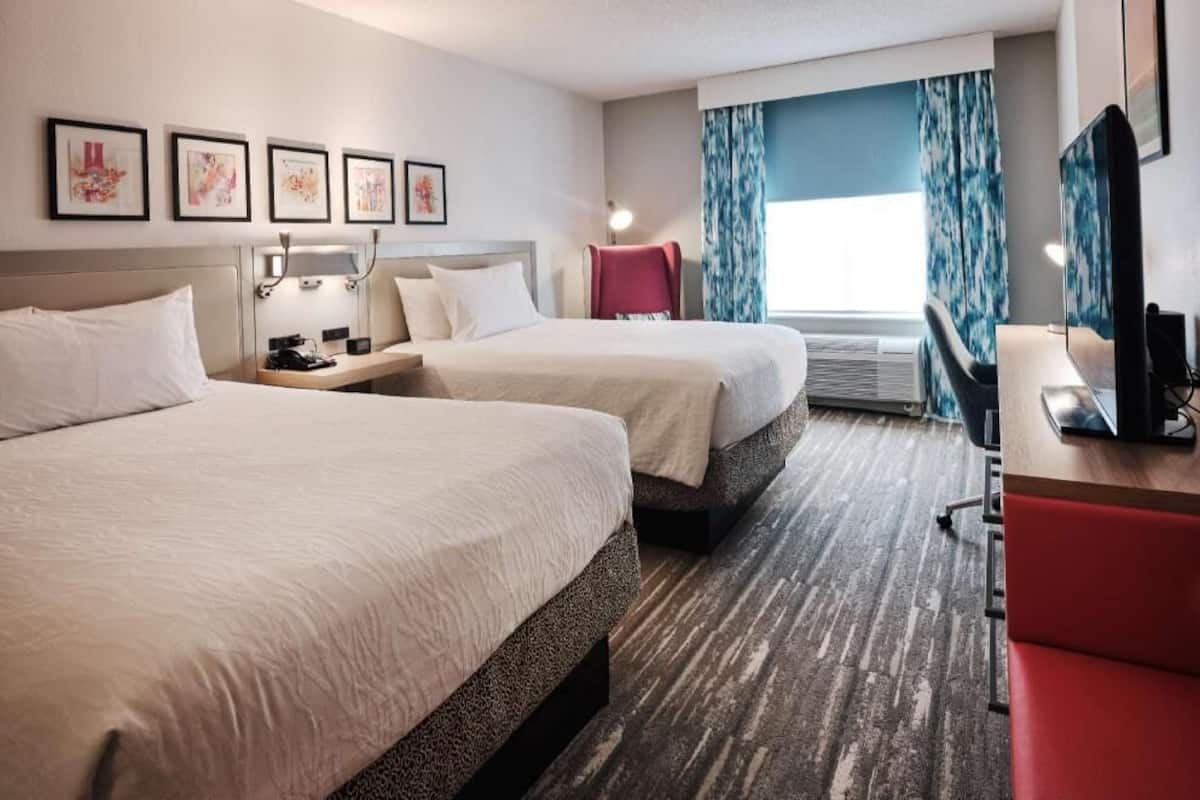 A hotel room with two beds, a desk, a chair, a TV, and a window with curtains.