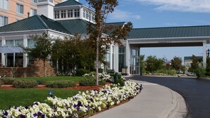 A hotel entrance with a covered driveway, a landscaped garden with white and purple flowers, and a building with a green roof and stone facade.