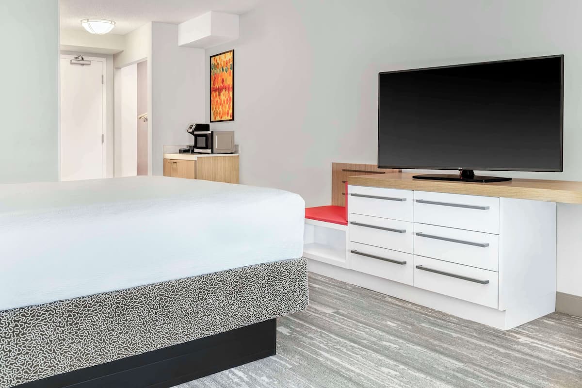 A modern hotel room with a bed, a flat-screen TV, a wooden side table, and a white dresser with drawers.