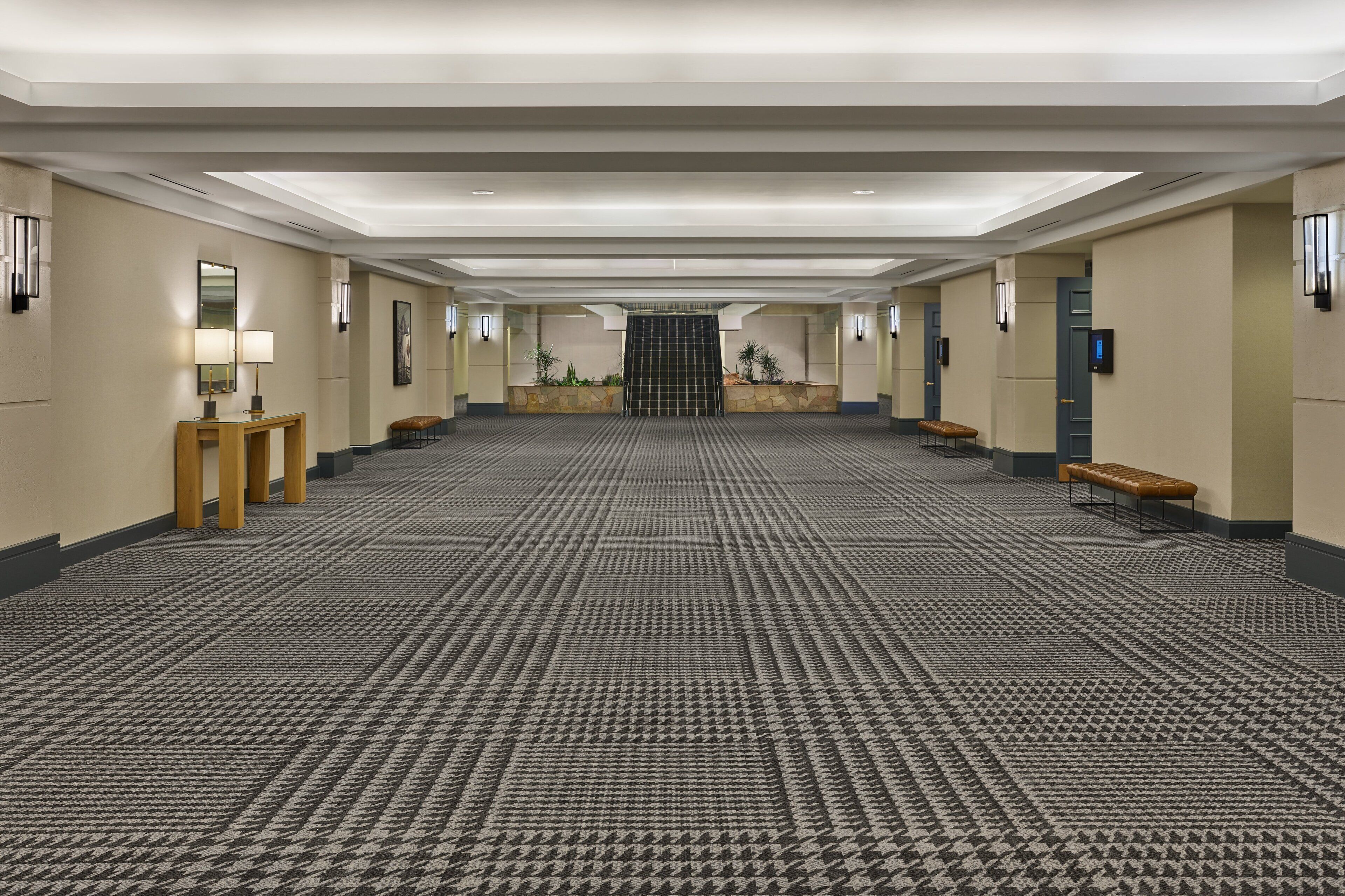 A spacious hotel lobby with a patterned carpet, wooden benches, a reception desk, and a staircase leading to an upper floor.