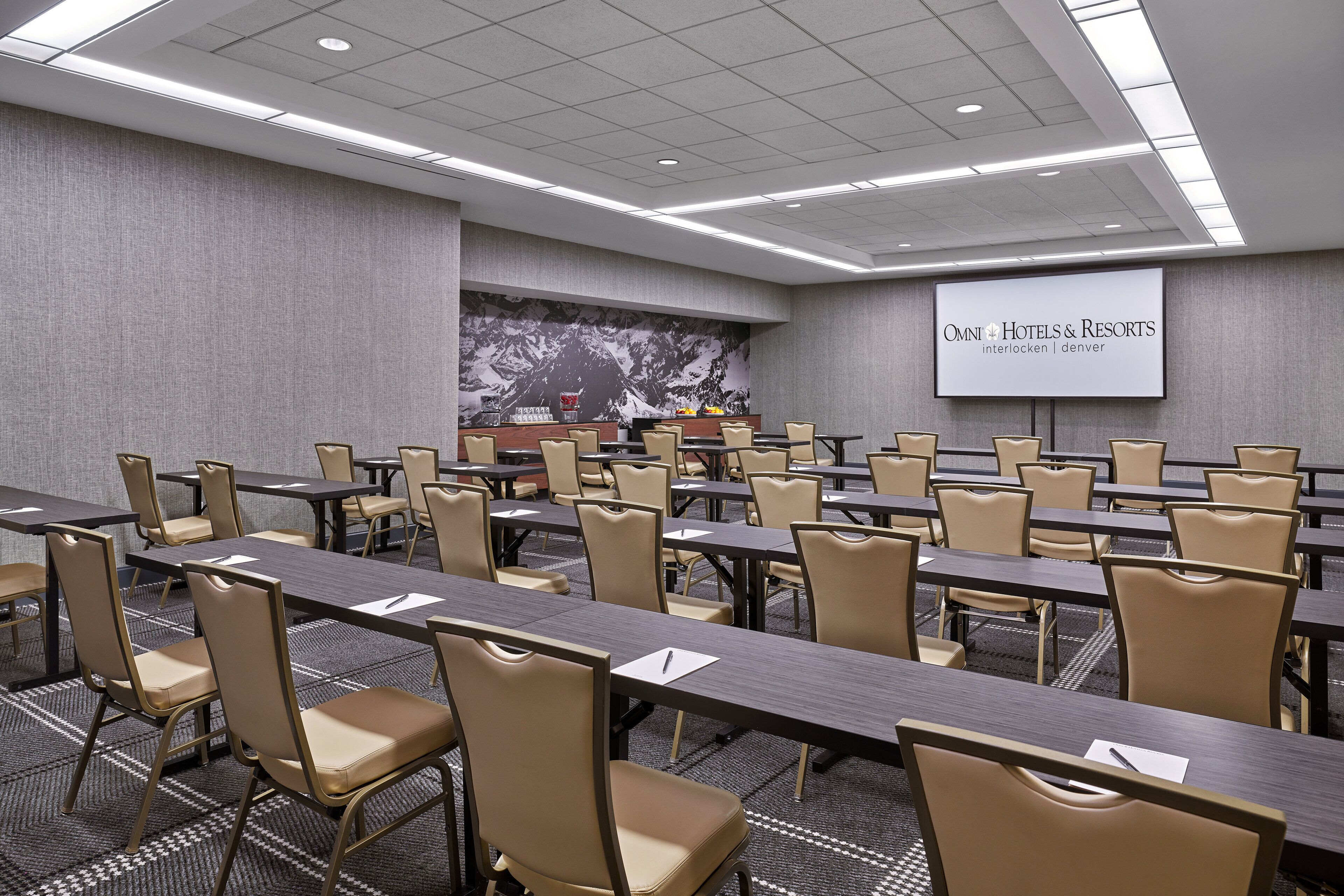 A conference room with multiple tables and chairs, a large screen displaying 'OMNI Hotels & Resorts Interlocken | Denver', and a mountain mural on the wall.