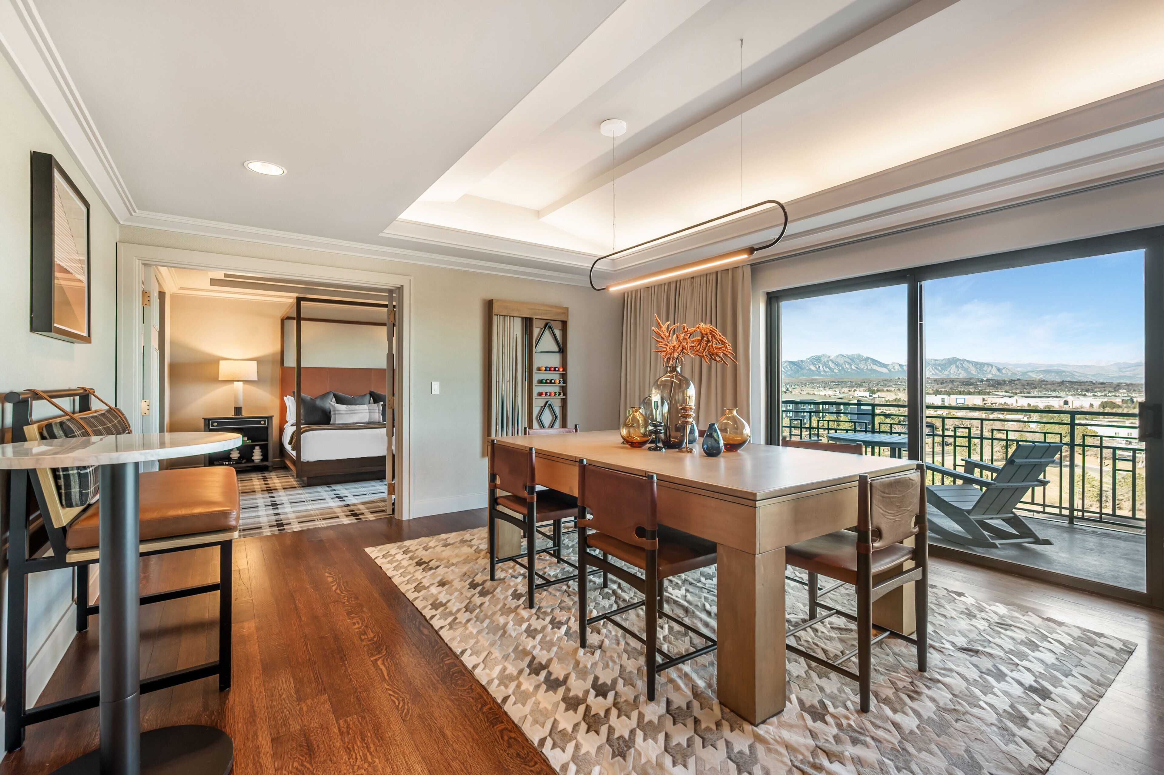 A modern hotel room with a dining area, a large window with a mountain view, and a balcony with outdoor seating.
