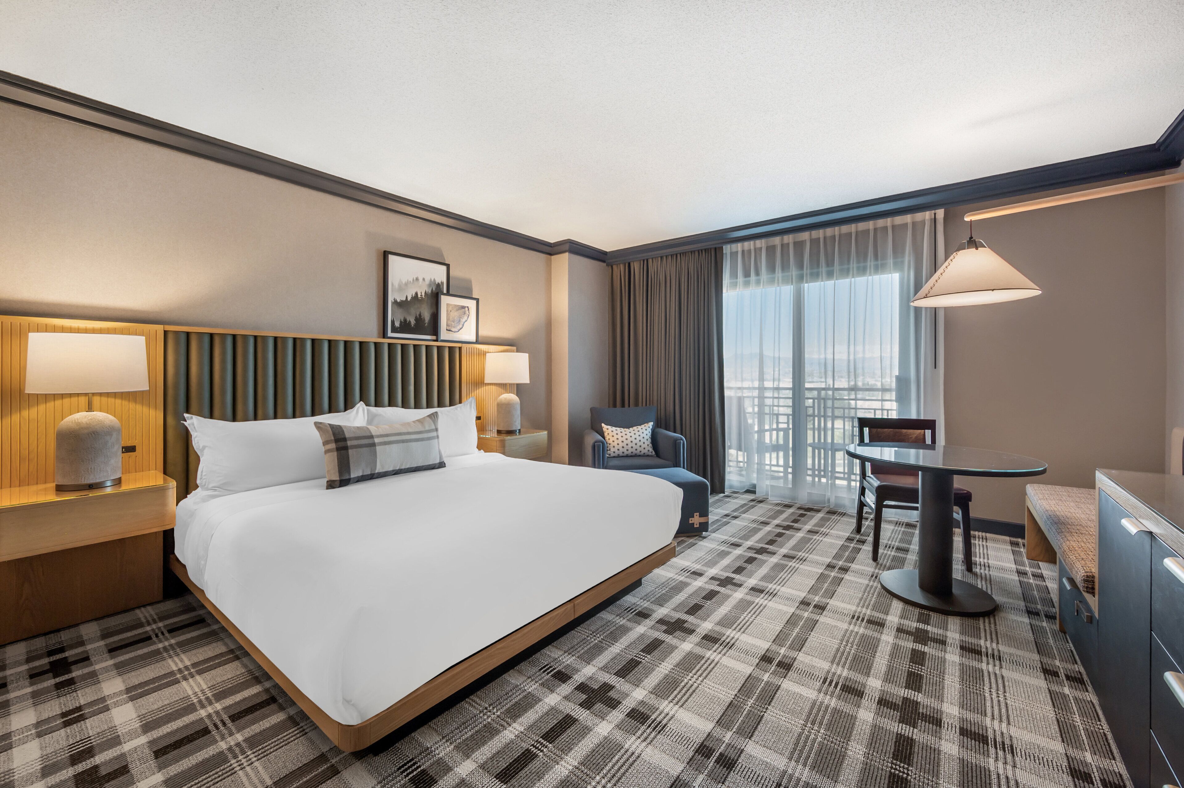 A hotel room with a large bed, a desk, a chair, a lamp, and a view of the city.