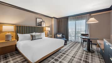 A hotel room with a large bed, a desk, a chair, a lamp, and a view of the city.
