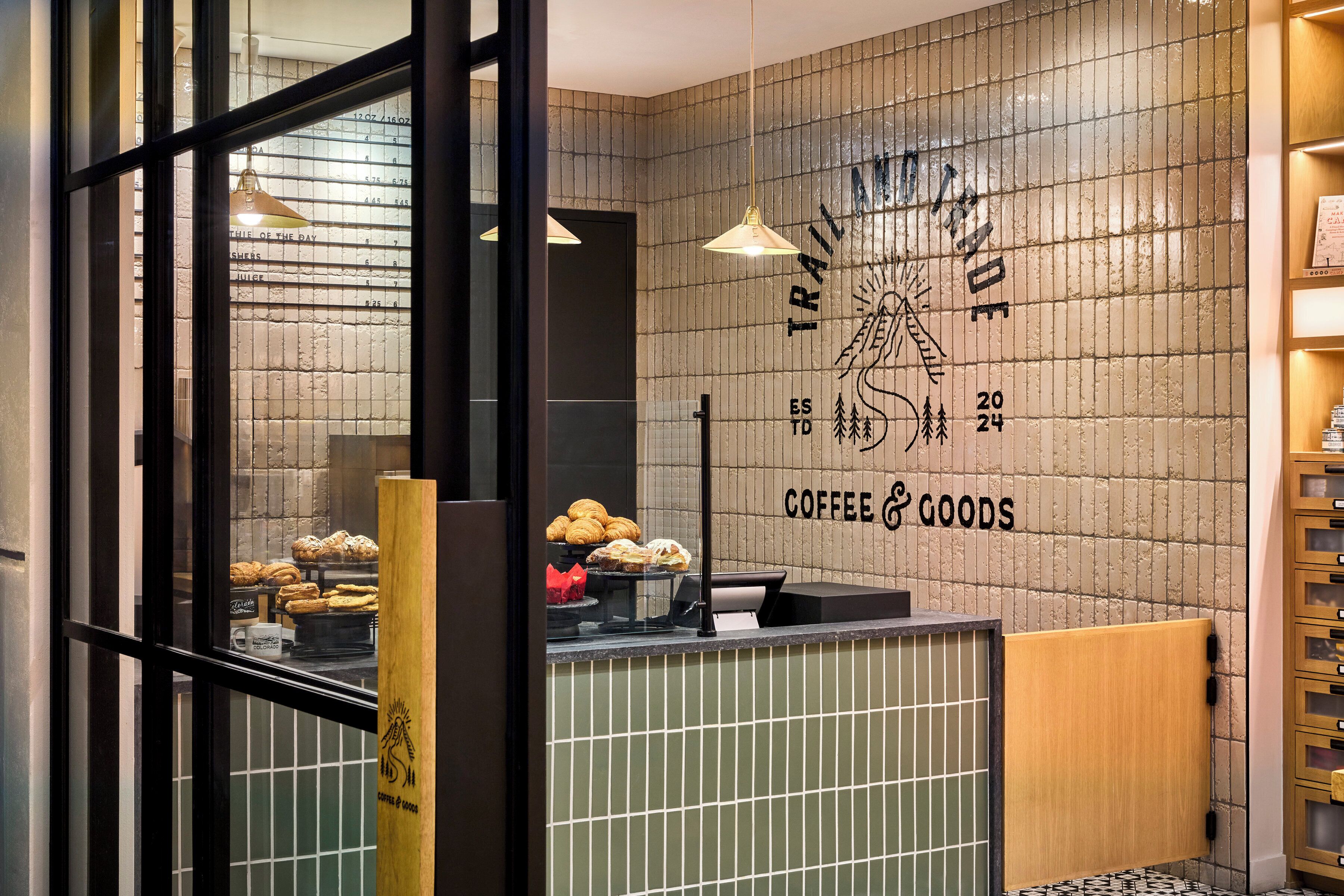 Interior d'una cafeteria amb un mostrador on es mostren pastissos, una paret amb un cartell de cafè i productes, i una prestatgeria de fusta amb articles.
