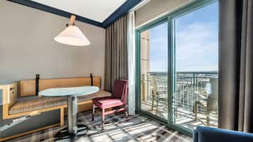 A hotel room with a large window, a round glass table, a red chair, a bench with a patterned cushion, and a hanging light fixture.