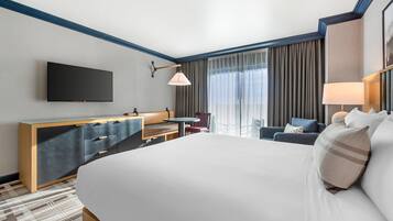 A hotel room with a large bed, a flat-screen TV mounted on the wall, a desk with a chair, and a window with curtains.