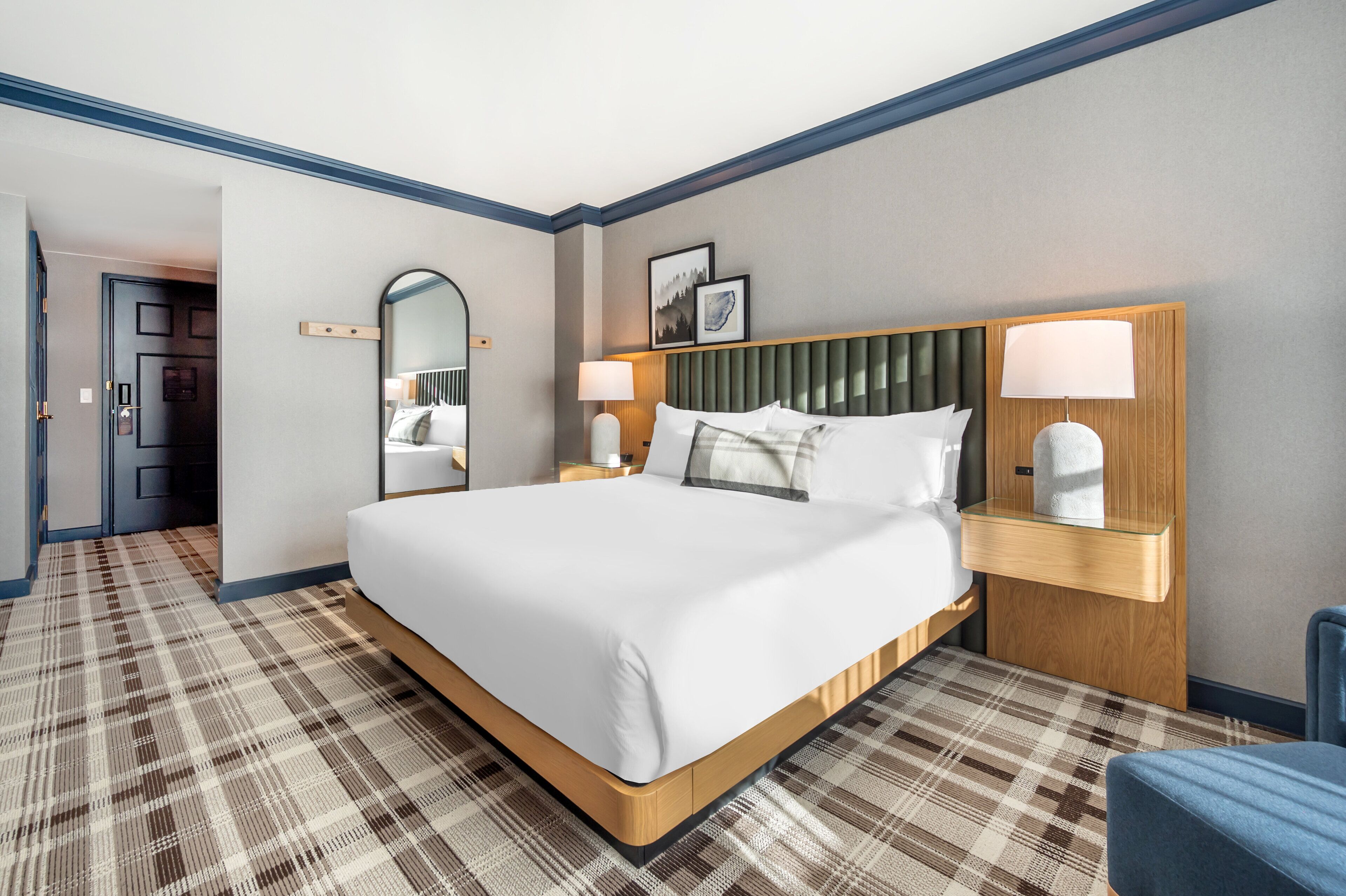 A hotel room with a large bed, a bedside table with lamps, a mirror, and a plaid-patterned carpet.