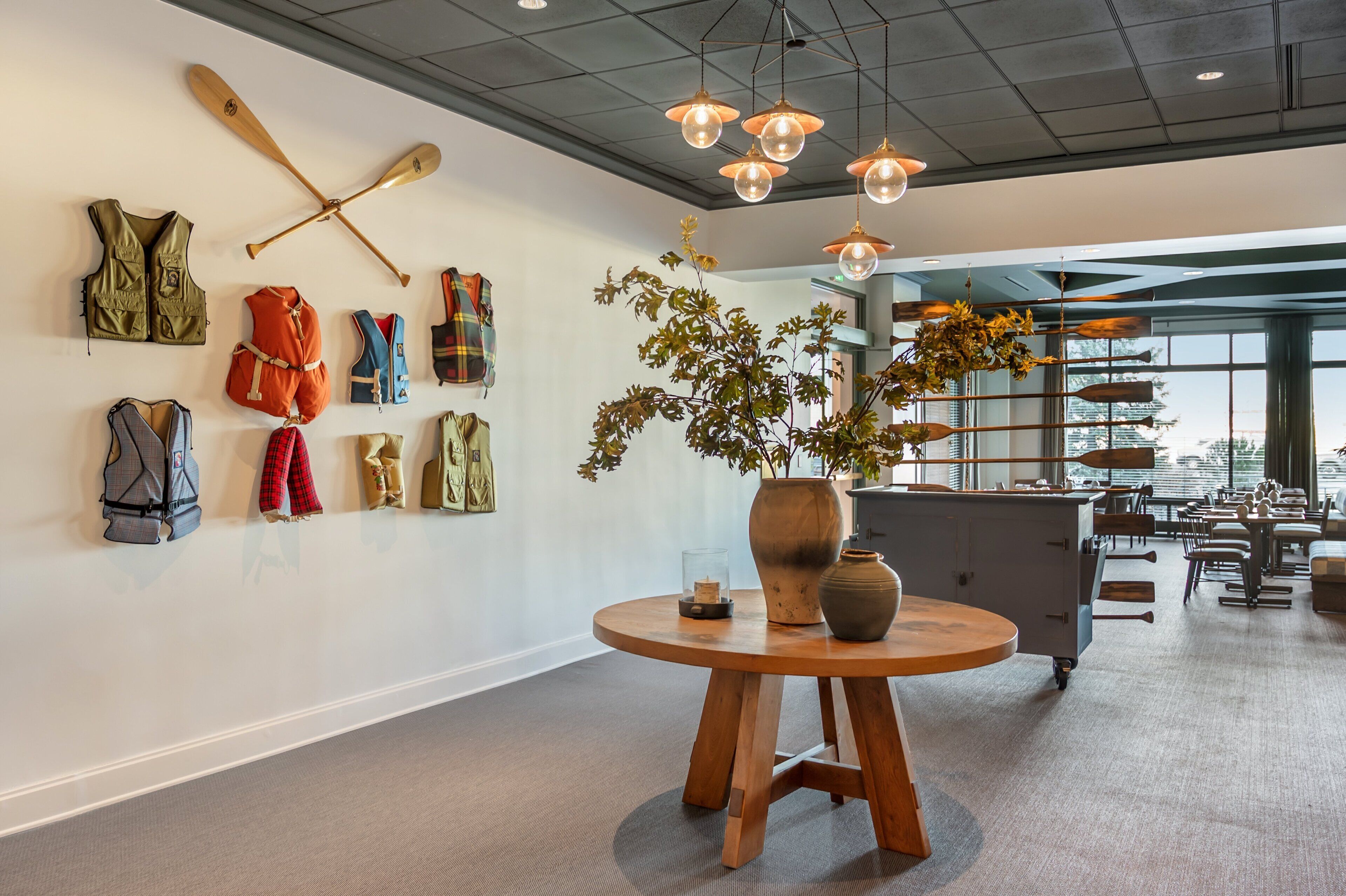 A modern interior with a wall displaying various outdoor gear, a wooden table with a vase and candle, and a view of the city through large windows.