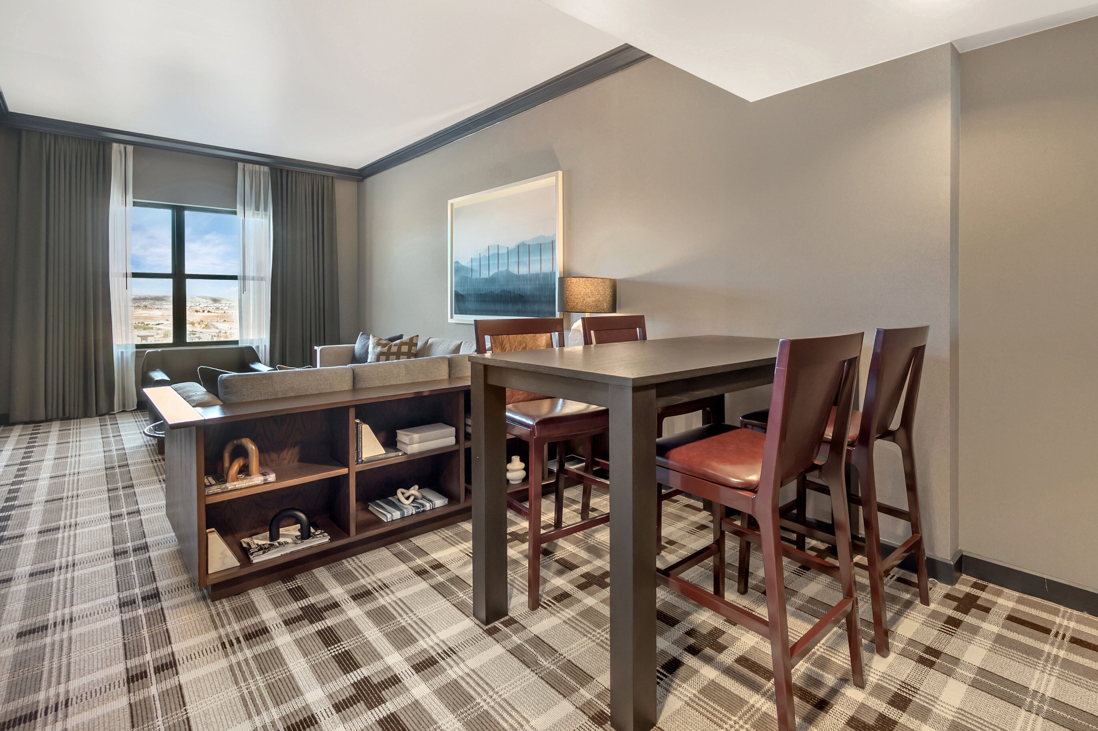A hotel room with a dining table, chairs, a sofa, a lamp, and a painting on the wall.