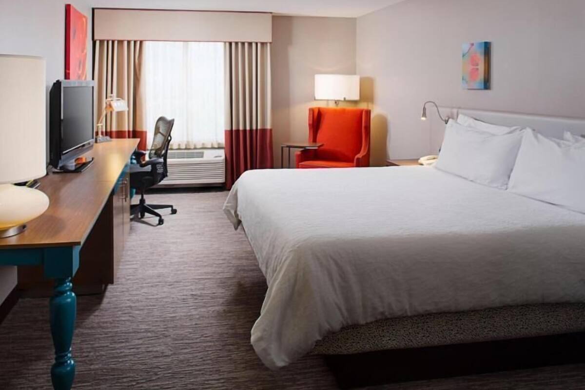A hotel room with a bed, a desk with a television, a chair, a lamp, and a window with curtains.