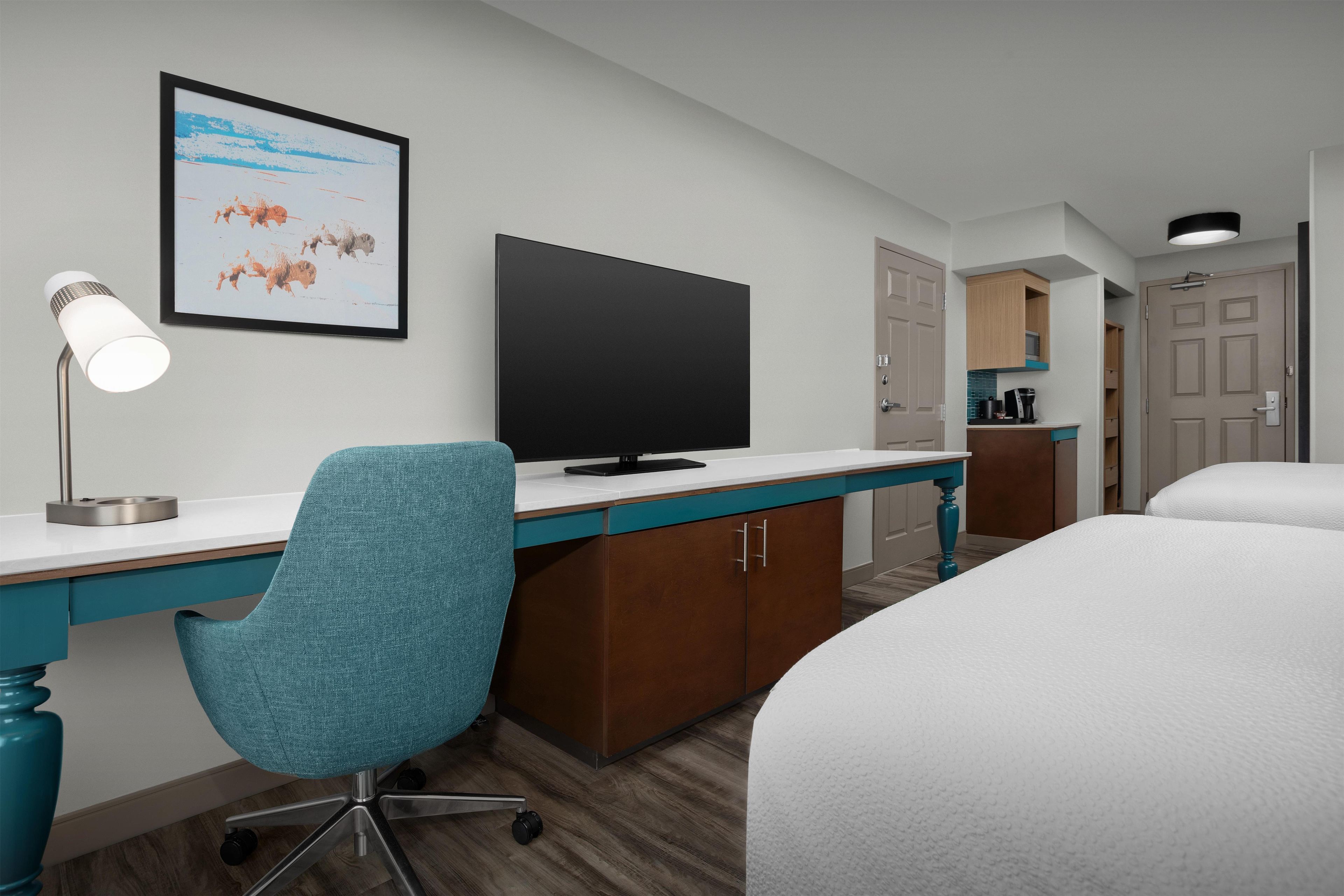 A hotel room with a bed, a desk with a computer, a chair, a TV, a lamp, and a framed picture on the wall.