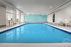 A large indoor swimming pool with a blue tiled edge, surrounded by white walls and columns, with seating areas and a door on the right. - Hilton Garden Inn Colorado Springs North Air Force Academy (Colorado Springs)