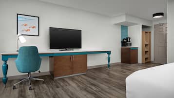 A hotel room with a wooden desk, teal chair, flat-screen TV, and a bed.