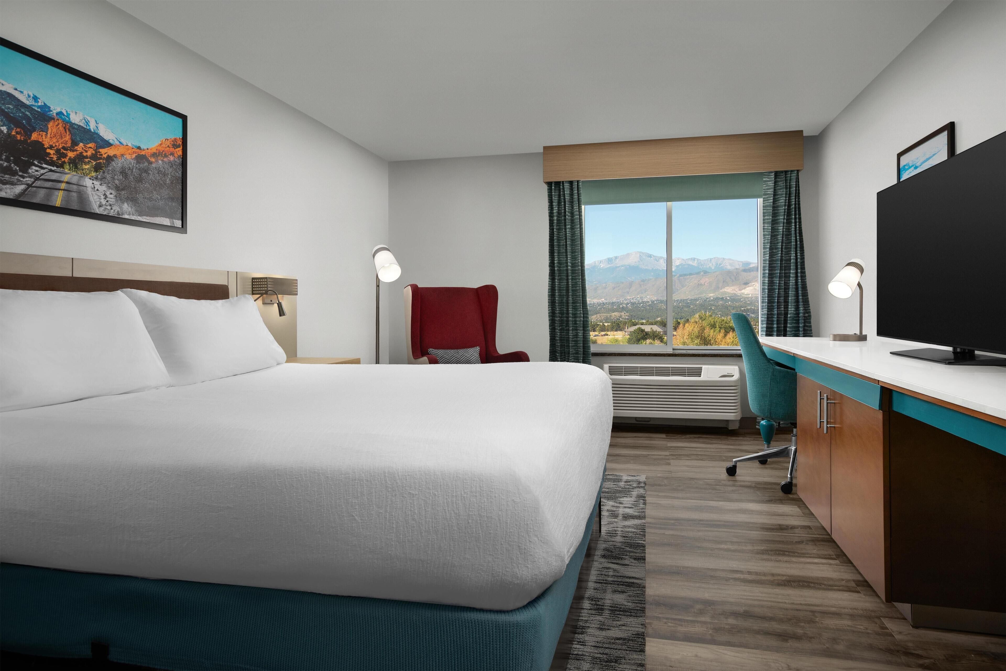 Habitación de hotel con cama, escritorio, silla, televisor y un amplio ventanal con vista a la montaña.