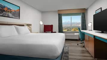 Habitación de hotel con cama, escritorio, silla, televisor y un amplio ventanal con vista a la montaña.