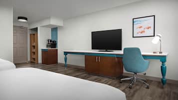 A hotel room with a bed, a desk with a flat-screen TV, a chair, and a desk lamp.
