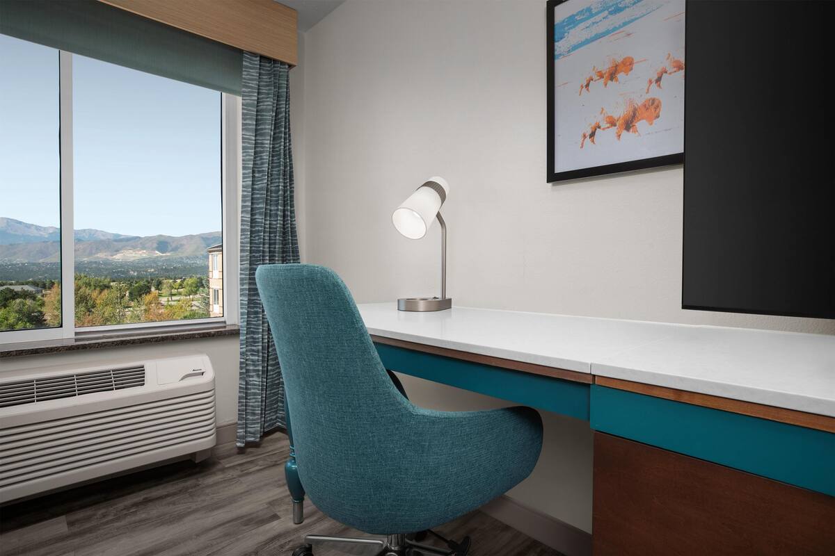 A modern office with a view of mountains, a desk, a chair, a lamp, and a framed picture.