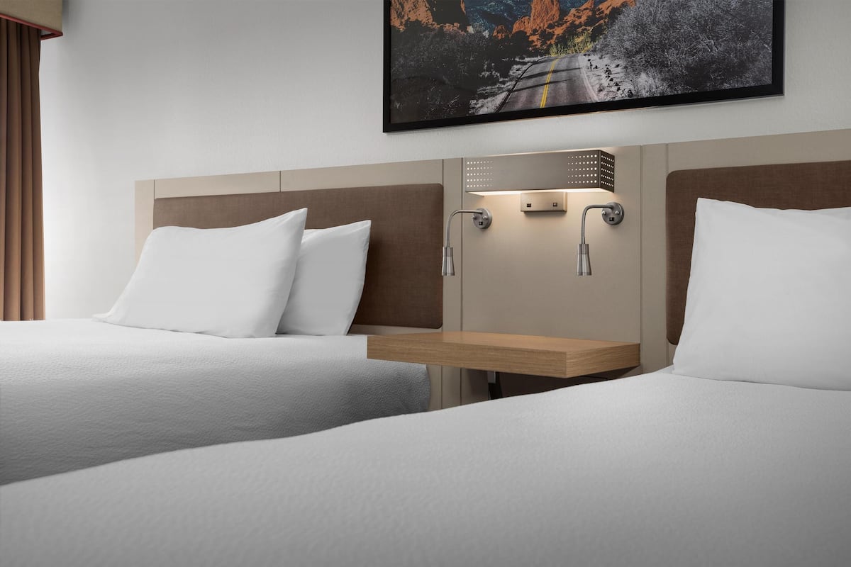 A hotel room with two beds, a nightstand, a wall-mounted light, and a painting above the bed.