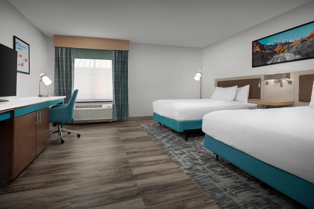 A hotel room with a desk, two beds, a chair, a window with curtains, a painting, and a wall-mounted heater.