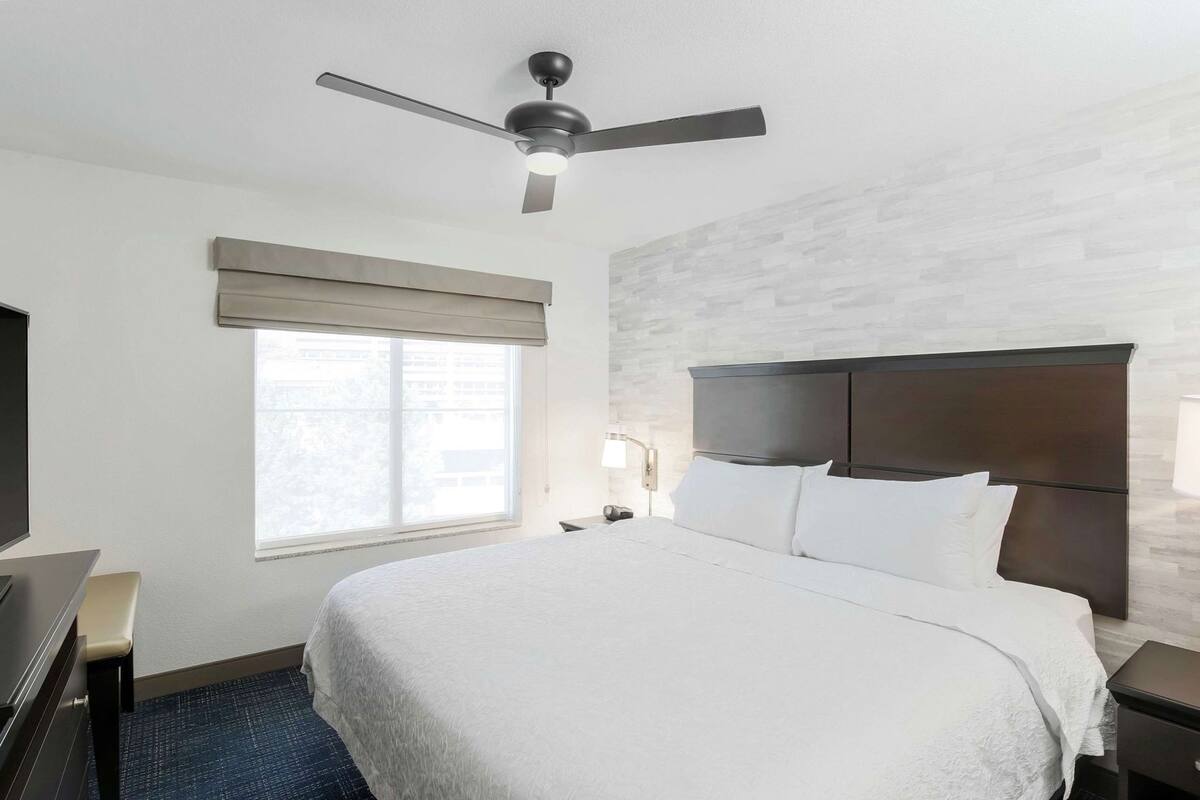 Una habitación de hotel con una cama, ventilador de techo, ventana con persianas, mesita de noche y lámpara.