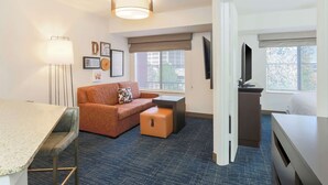 Une chambre d’hôtel équipée d’une kitchenette, d’un canapé, d’une petite table, d’une télévision et d’une fenêtre donnant sur des immeubles.