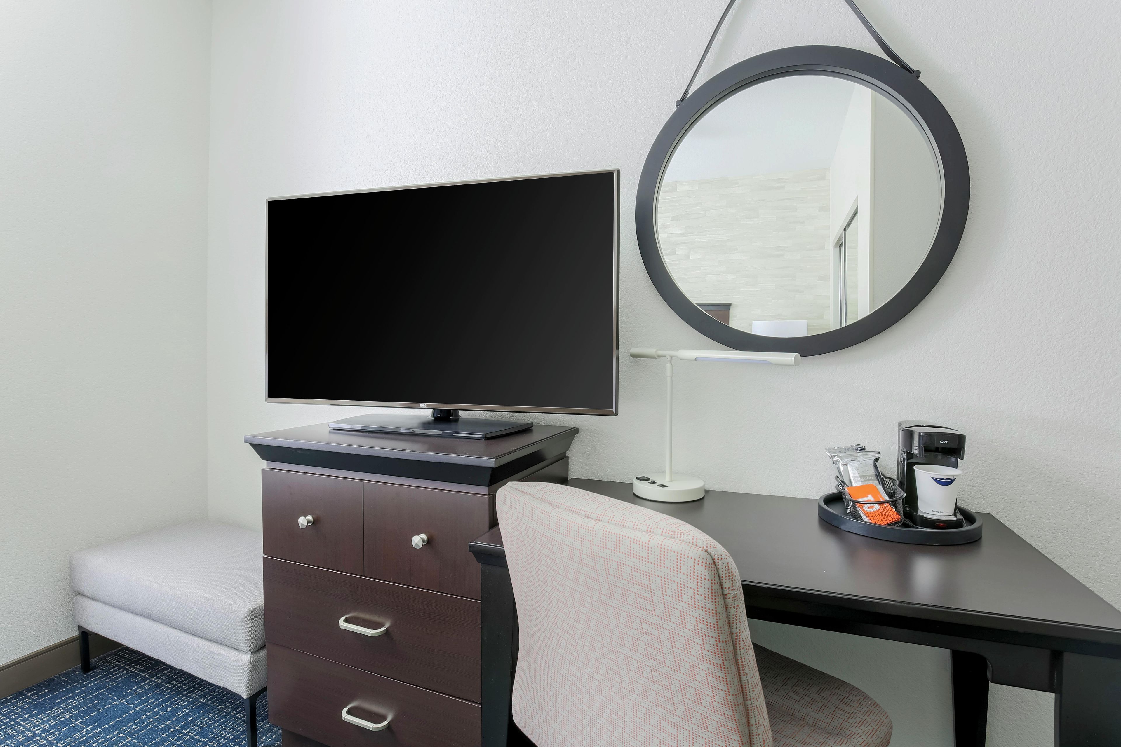 Ein Hotelzimmer mit Fernseher auf einer Kommode, einem runden Spiegel an der Wand, einem kleinen Tisch mit Kaffeemaschine und einem gepolsterten Sessel.