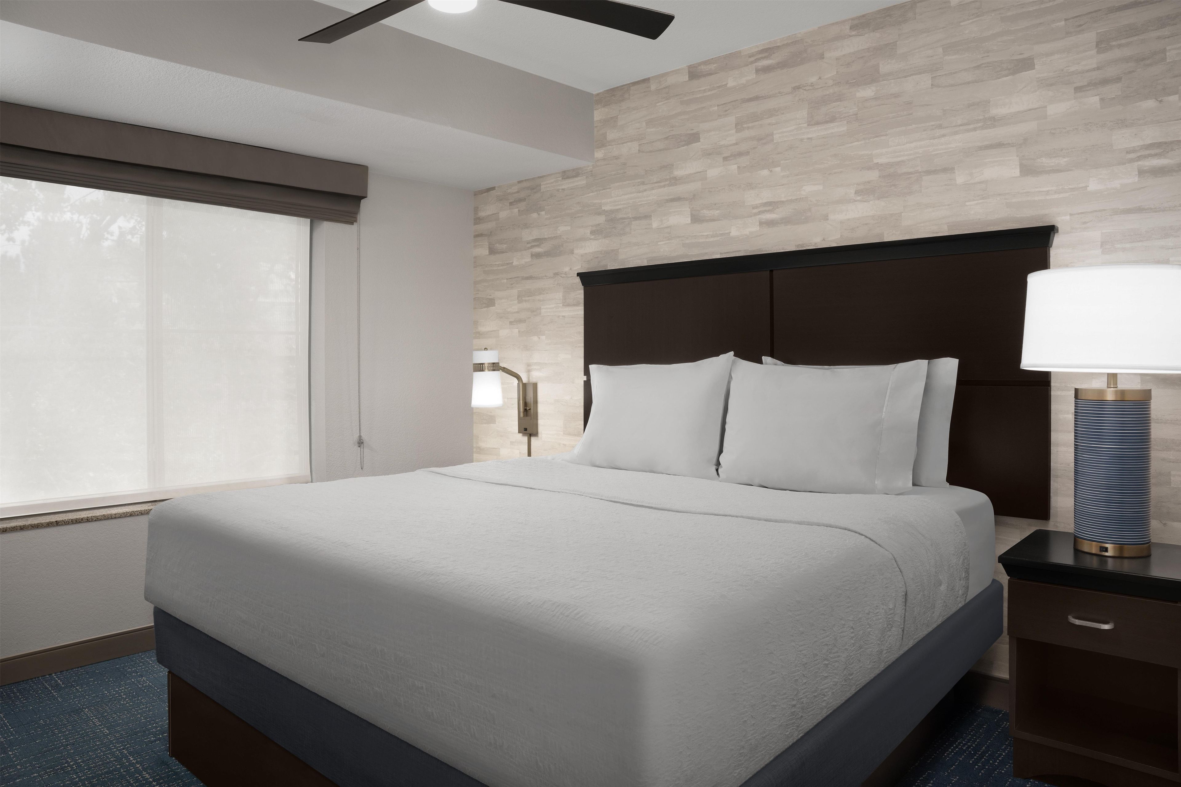 A hotel room with a bed, a nightstand, a lamp, and a window with blinds.