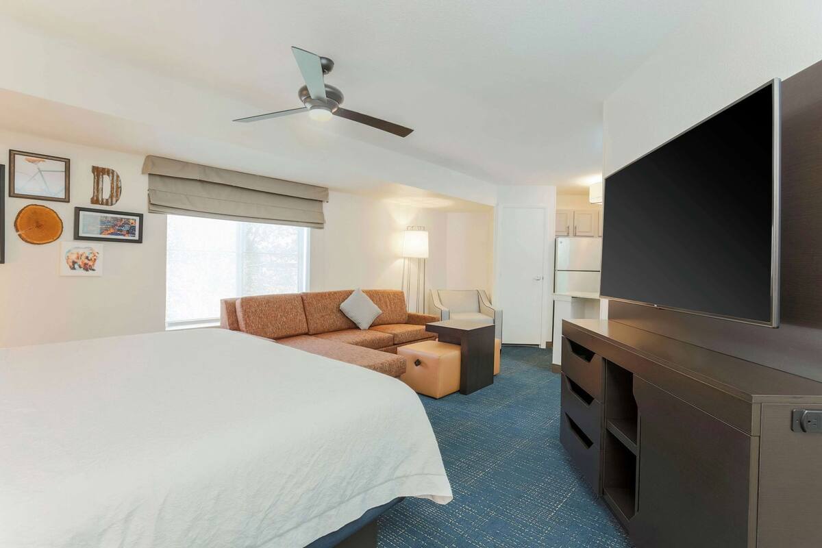 Una habitación de hotel con cama, sofá, televisión, ventilador de techo y ventana con persianas.