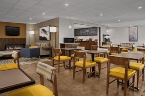 A modern restaurant with wooden tables and yellow chairs, a fireplace, and a kitchen area. - Hampton Inn & Suites Denver Tech Center (Denver)
