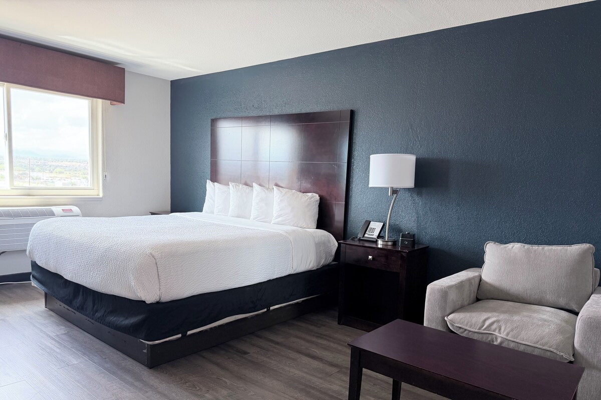 A hotel room with a bed, bedside table, lamp, chair, and a view of the city.
