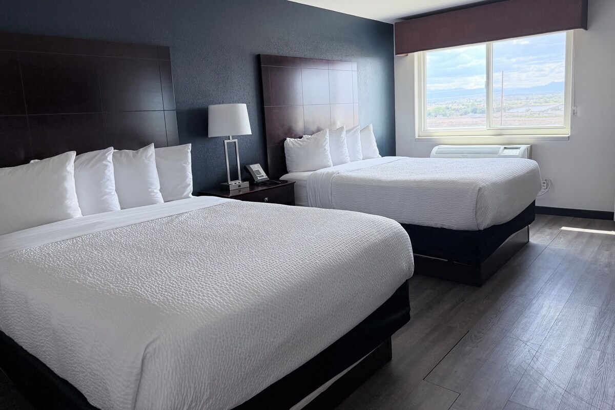 A hotel room with two beds, a nightstand with a lamp, and a window with a city view.