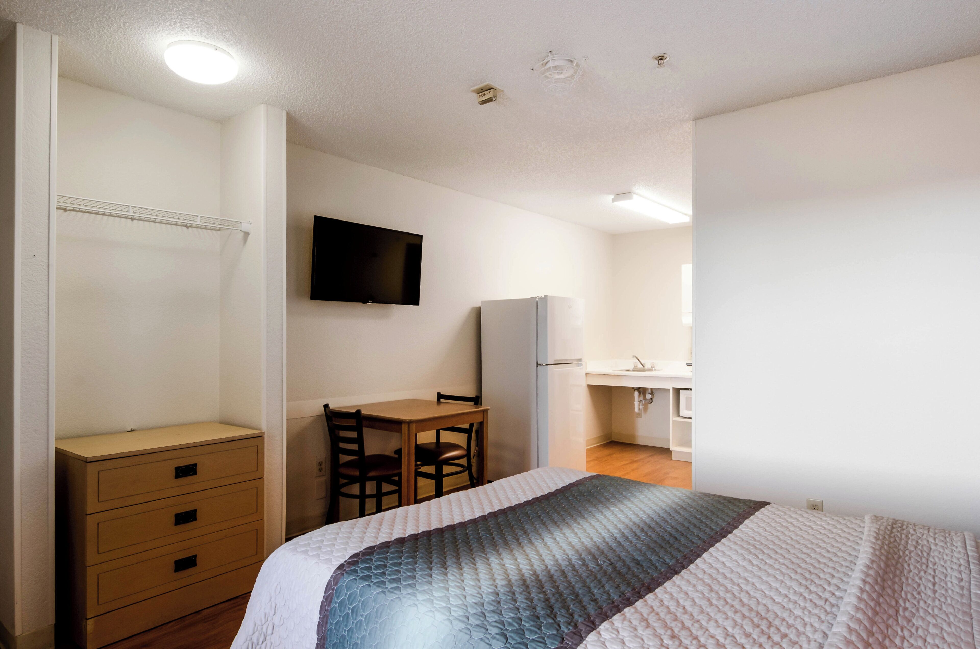 Ein Schlafzimmer mit Bett, Schreibtisch, Stuhl, Fernseher und einer Küchenzeile.
