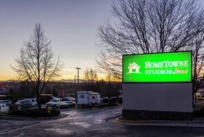 Naamplaatje voor Home Towne Studios by RedRoof in een parkeerplaats, met bomen en een zonsondergang op de achtergrond.