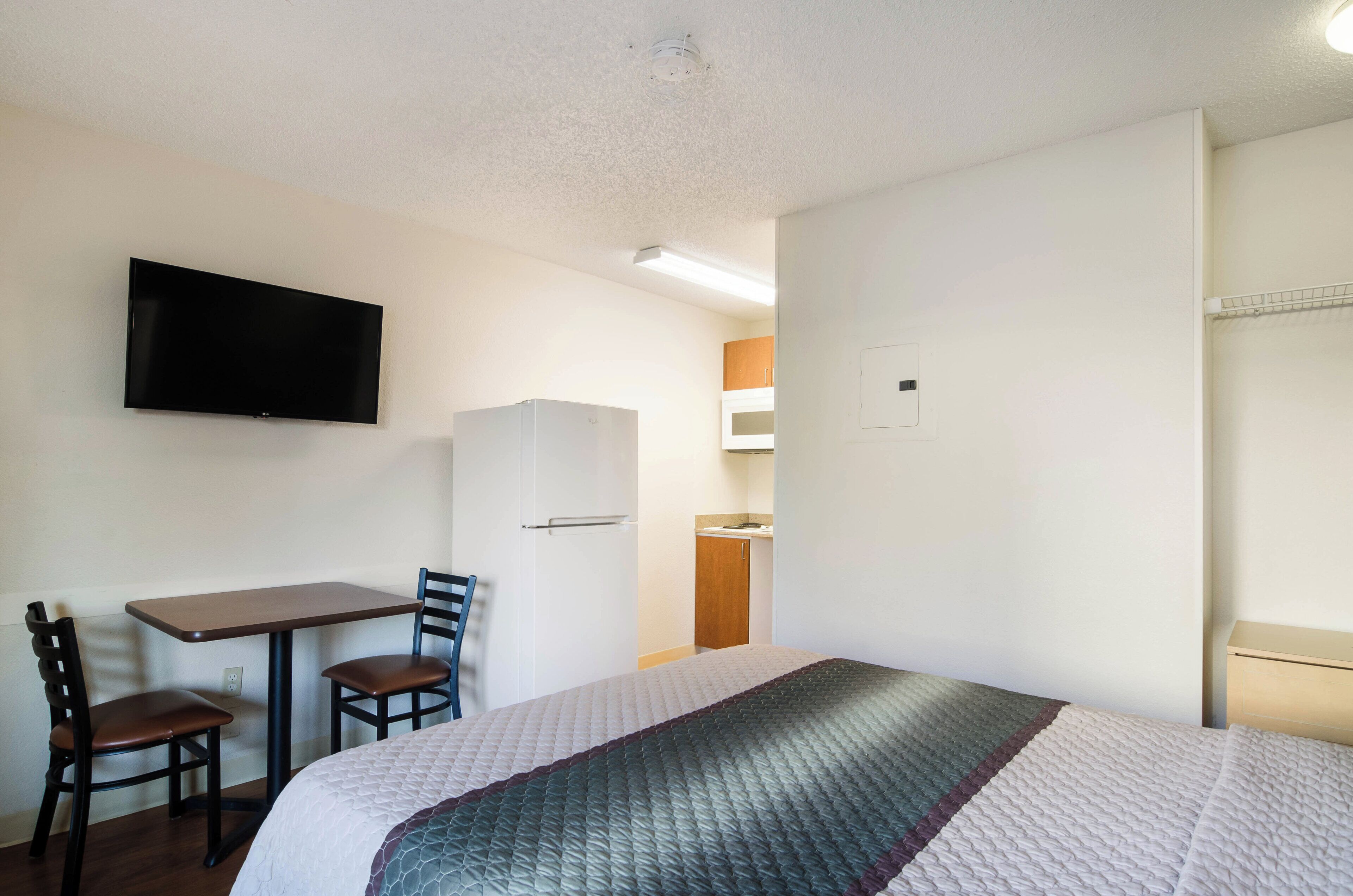 Una habitación de hotel con cama, escritorio con silla, televisor montado en la pared y una zona de kitchenette.
