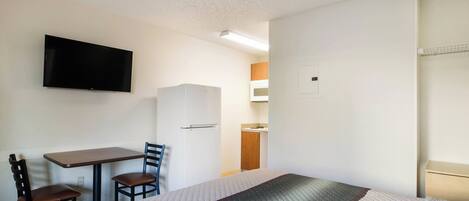 A hotel room with a bed, a desk with a chair, a television mounted on the wall, and a kitchenette area.