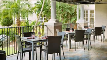 An outdoor patio with tables and chairs, surrounded by lush greenery and white columns.