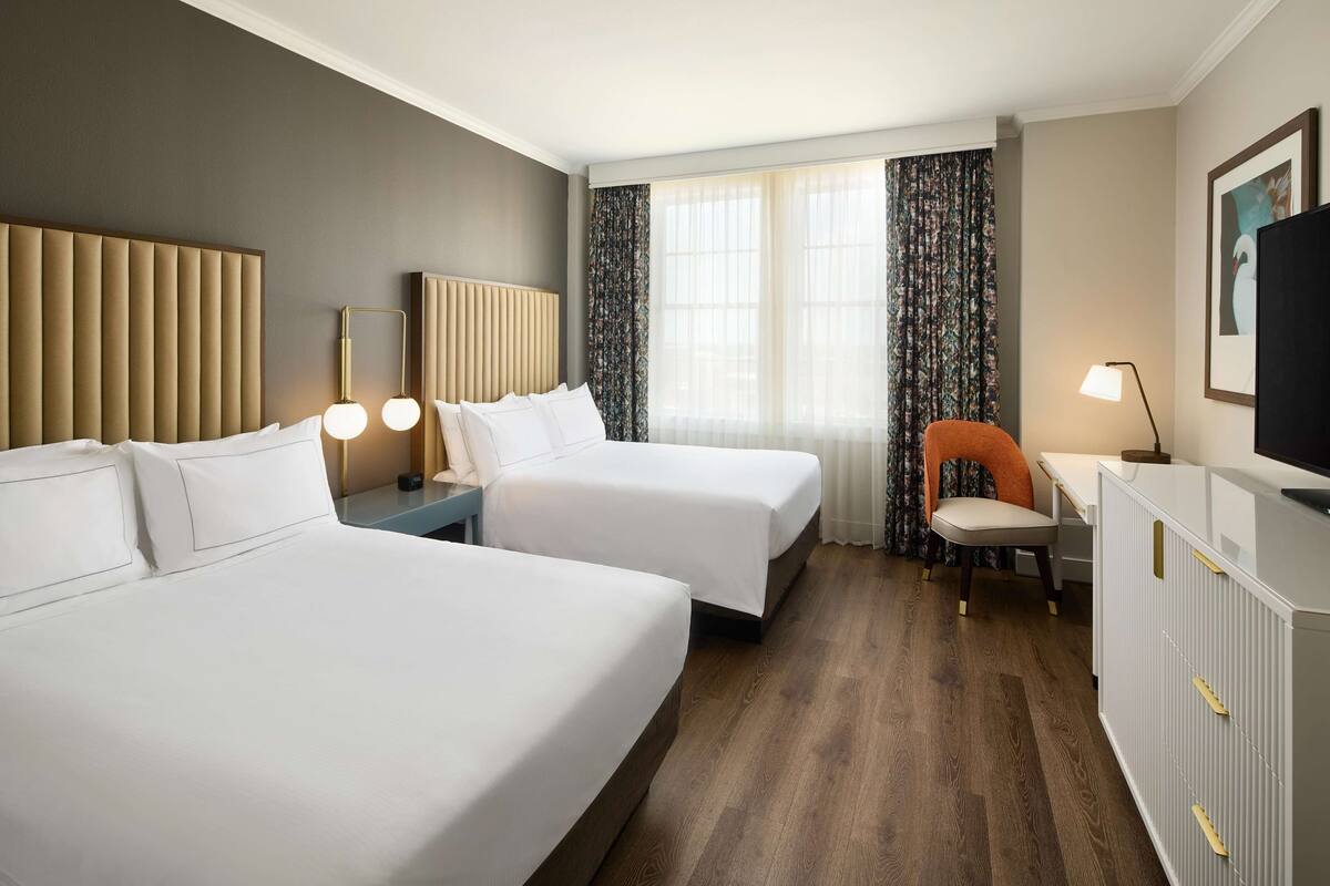 Habitación de hotel con dos camas, un escritorio, una silla, un televisor y una ventana con cortinas.
