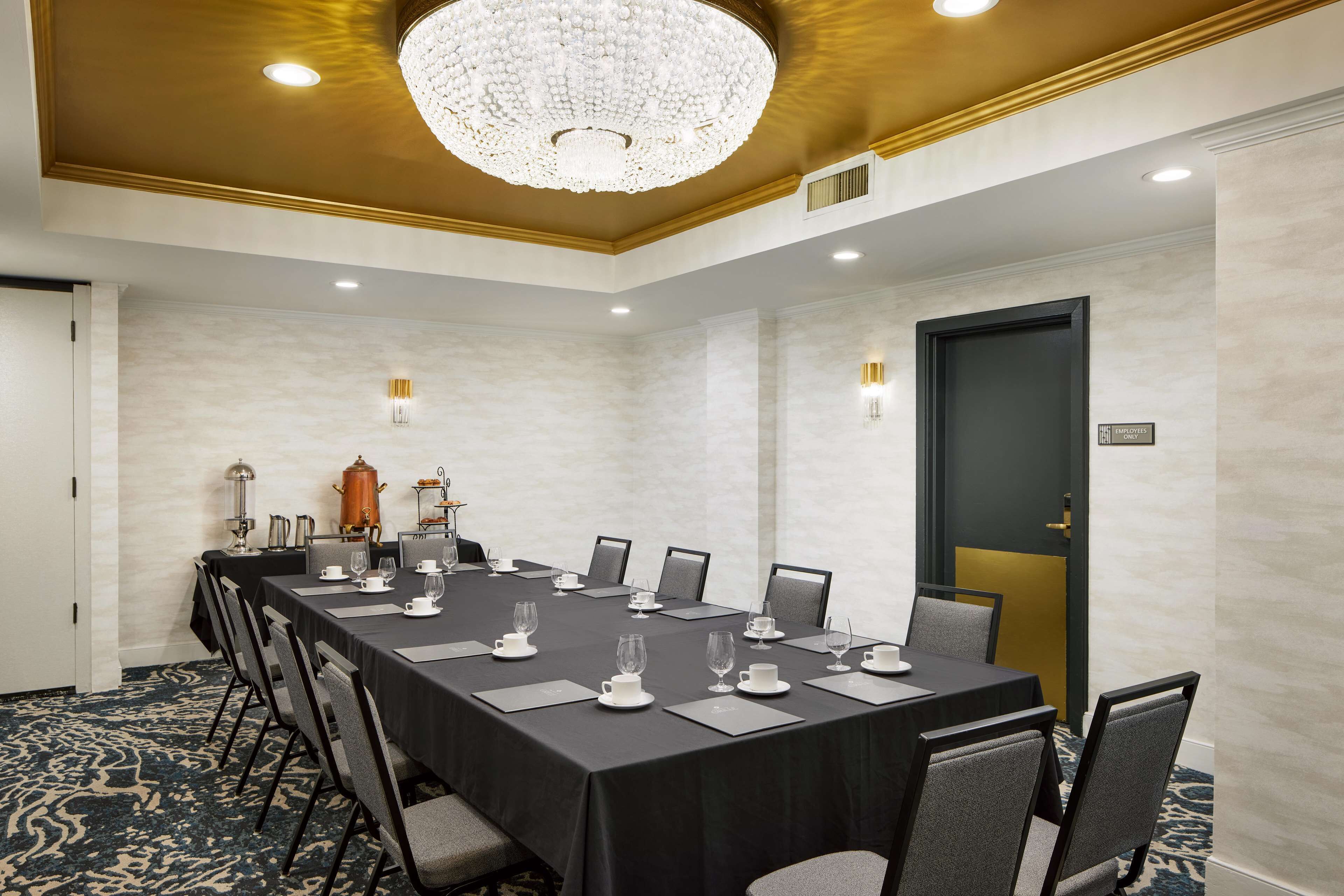 A conference room with a long table set for a meeting, equipped with chairs, a chandelier, and a coffee station.