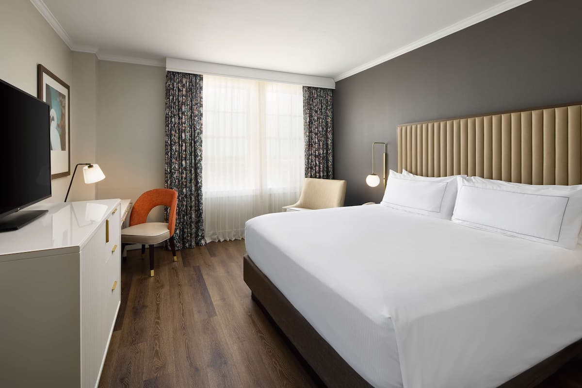 Habitación de hotel con una cama grande, un escritorio con televisor de pantalla plana, una silla y una ventana con cortinas.