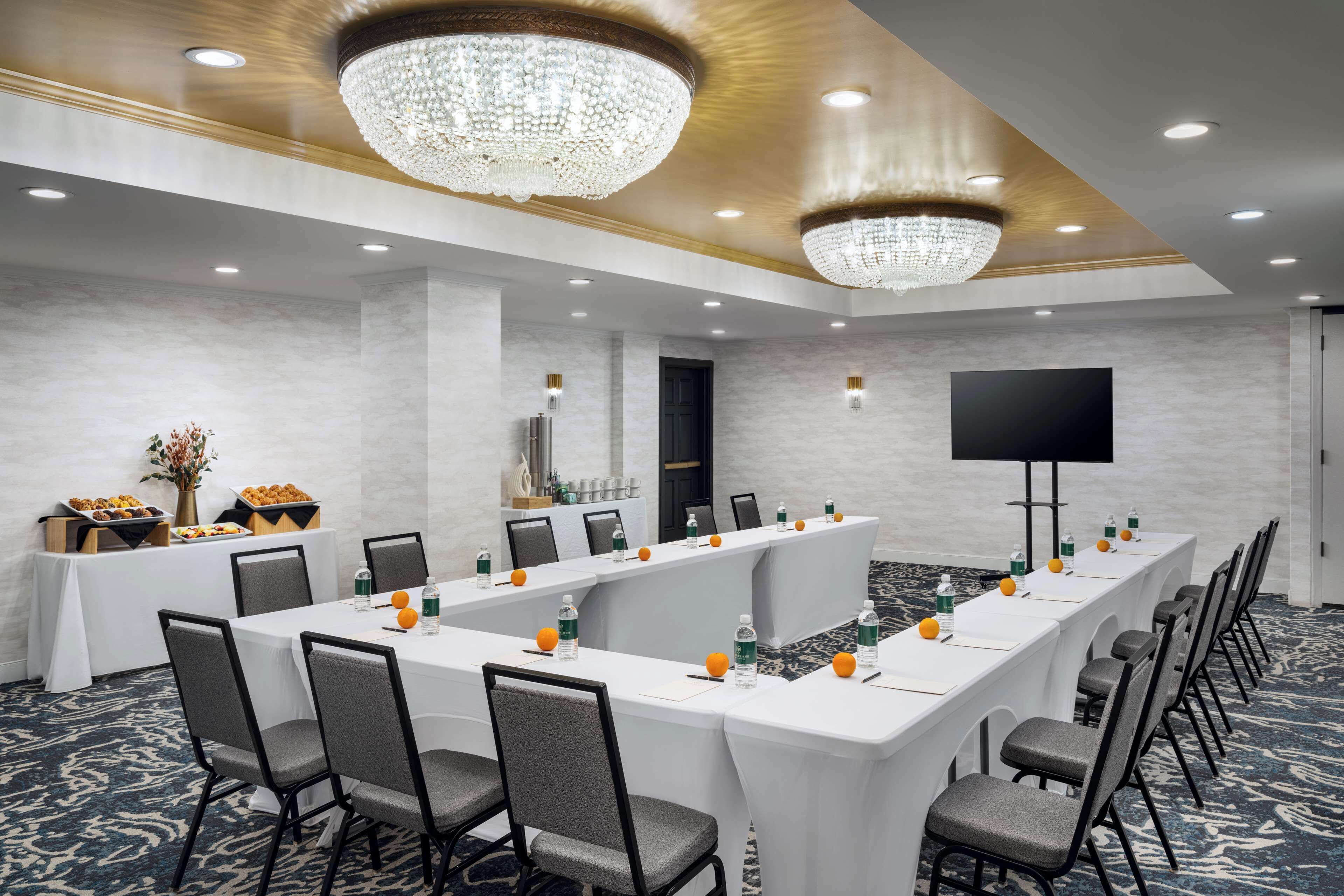 A conference room with a long table, chairs, a chandelier, a flat-screen TV, and a buffet area with food and drinks.