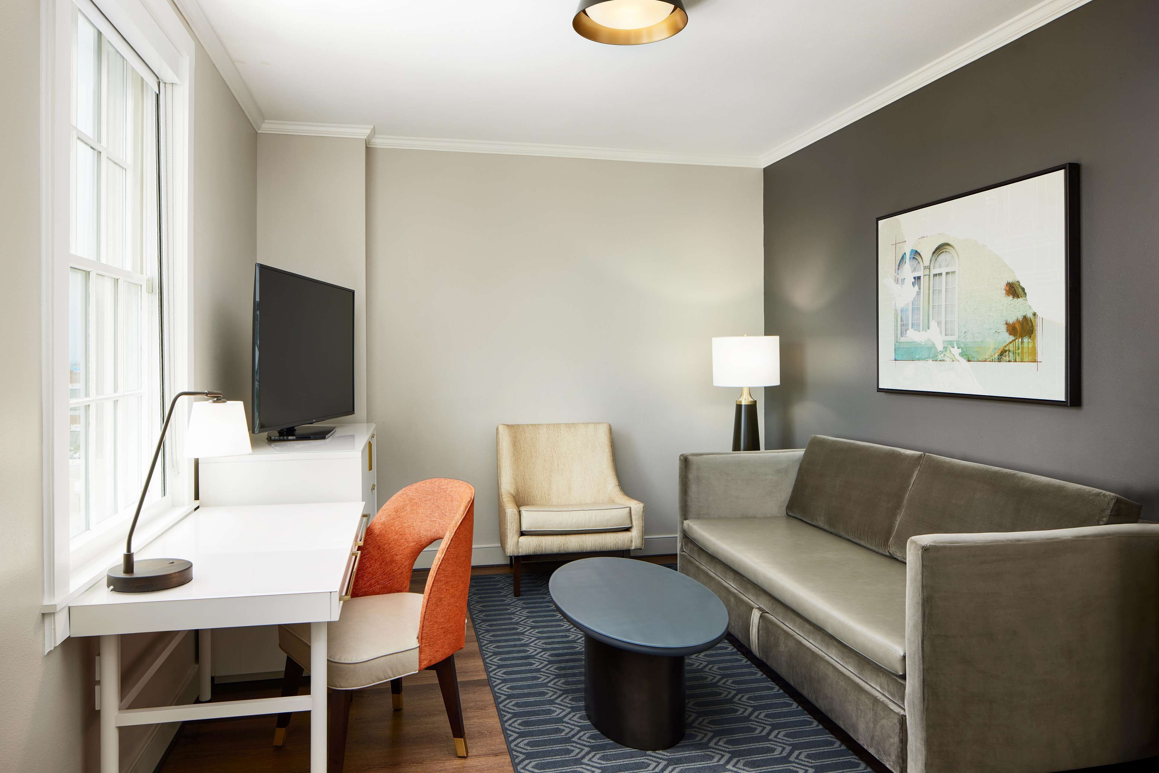A modern hotel room with a white desk, a television, a beige armchair, a grey sofa, and a patterned rug.